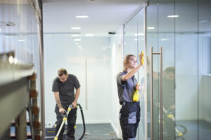 Office Cleaning Company Peoria IL, a female cleaning contractor is polishing the glass partition offices whilst In the background a male colleague steam cleans an office carpet in a empty office in between tenants. .
